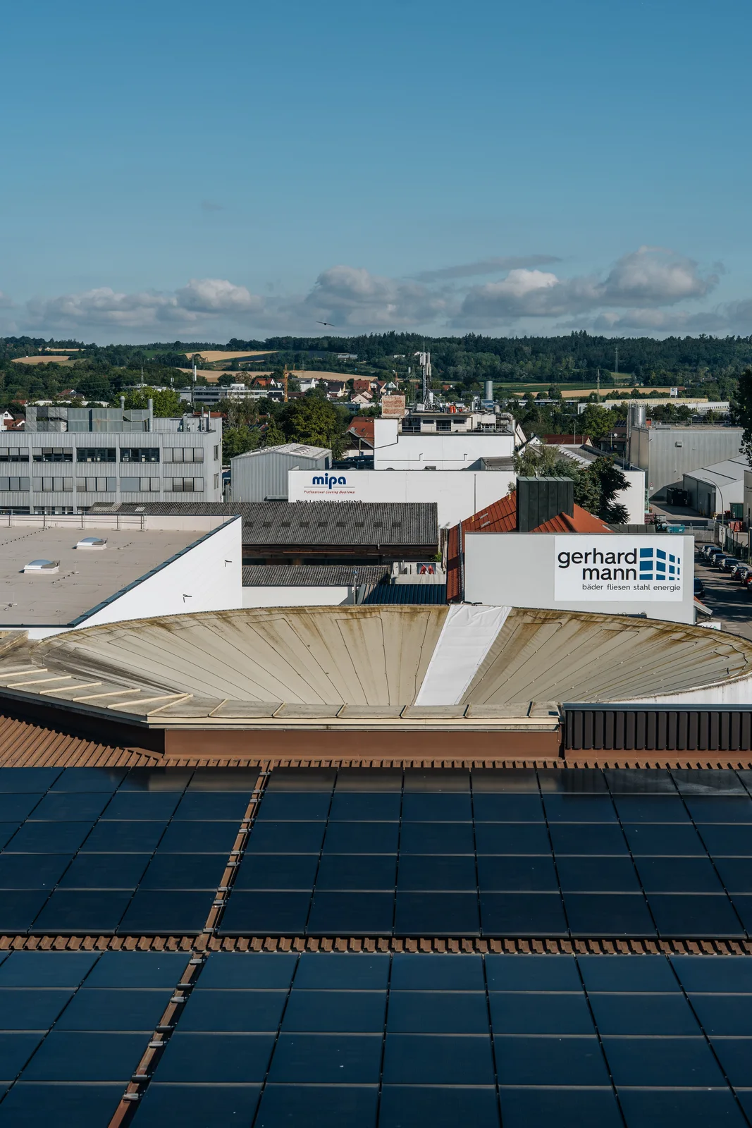 Photovoltaikmodule auf dem Dach, dahinter das Gerhard-Mann-Firmenschild und Hügel am Horizont