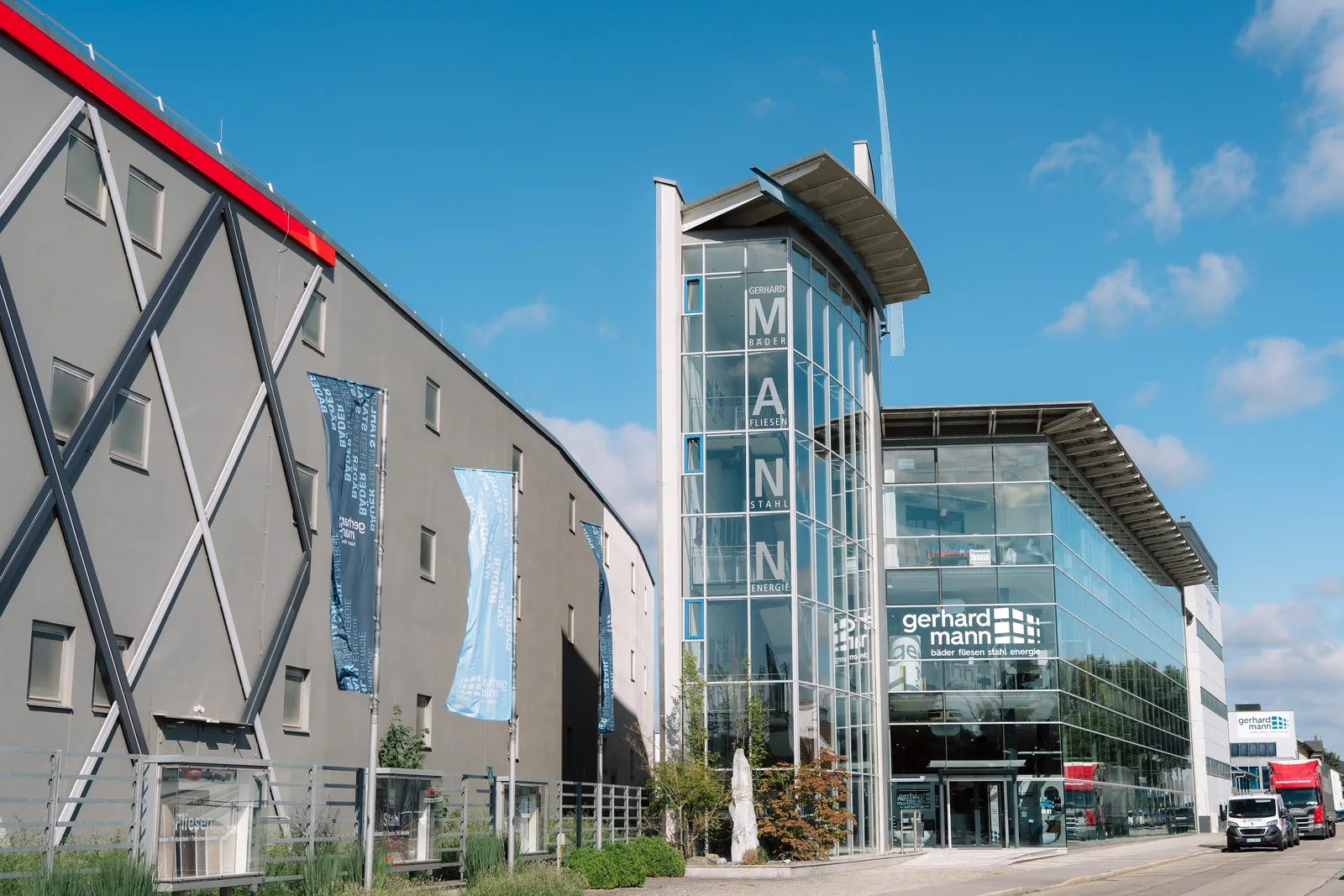 Straßenansicht des Gerhard-Mann-Standorts mit Halle, Fahnen, gläsernem Treppenturm und Glasfassade mit Firmenschriftzug